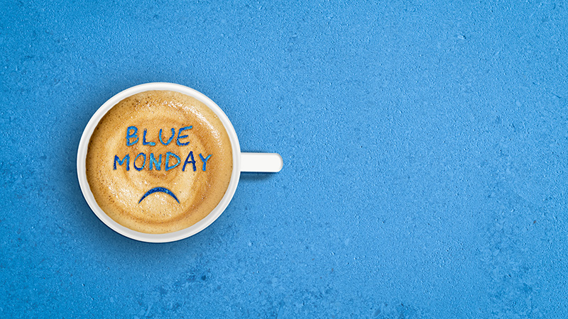 Image with a white cup of frothy coffee. Words BLUE MONDAY and sad mouth drawn in the layer of foam. Top view. Blue textured background. Blue Monday, the Monday of the last full week of January is the day when most people feel sad, down, or melancholy.