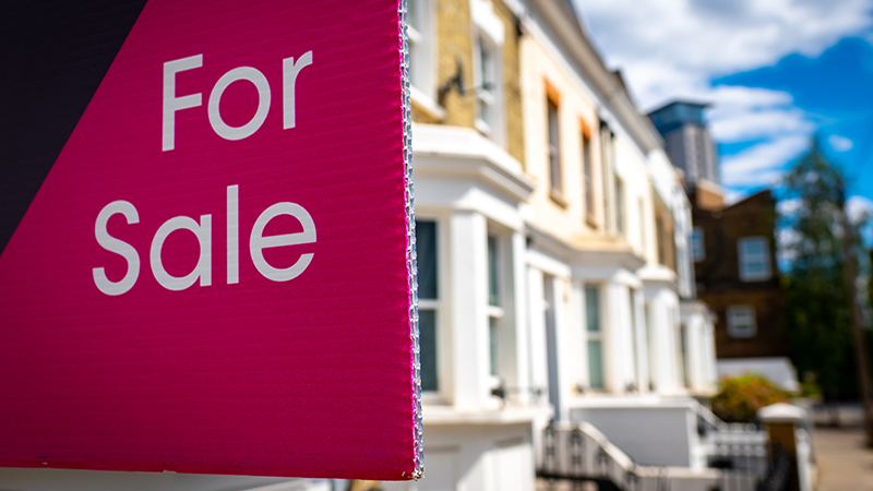 Red estate agent sign outside residential street of houses with 'For Sale' text
