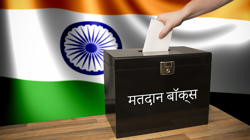 A hand casting a vote in a black ballot box for an election in India. The text says "ballot box" in Hindi.