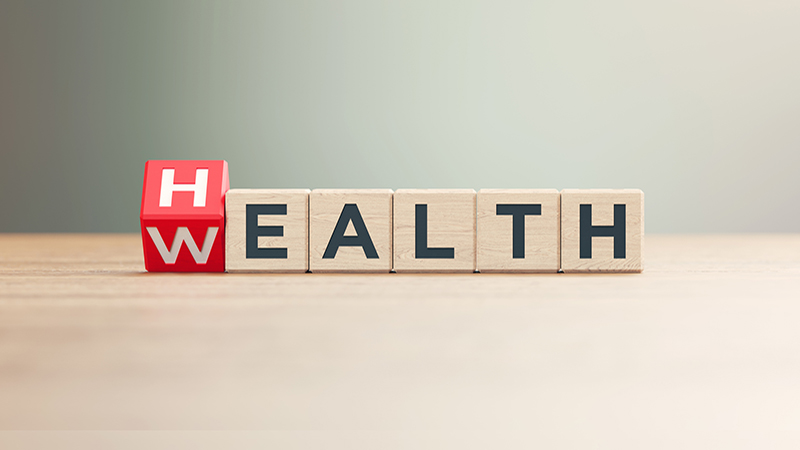 "Wealth" and "Health" written wood blocks sitting on a wood surface in front of a defocused background. Horizontal composition with copy space.