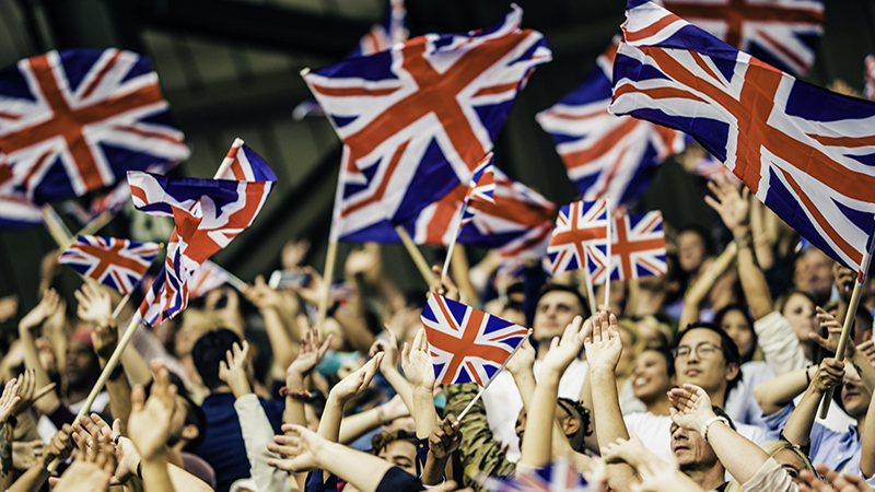 Large crowd in a football stadium cheering for their team with their hands raised and waving Great Britain flags.