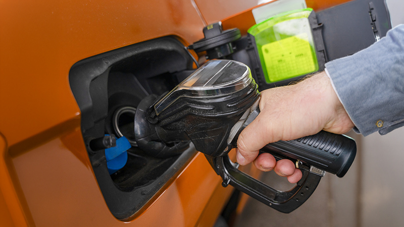 A person is fueling their vehicle at a gas station. They hold the pump and insert it into the gas tank. The scene shows the bright orange vehicle.