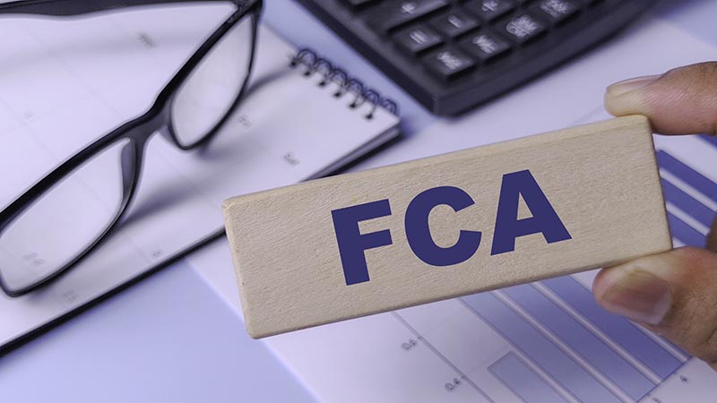 FCA (Financial Conduct Authority) Concept: Wooden Block with Abbreviation Held Over Financial Chart, Calculator, and Notebook